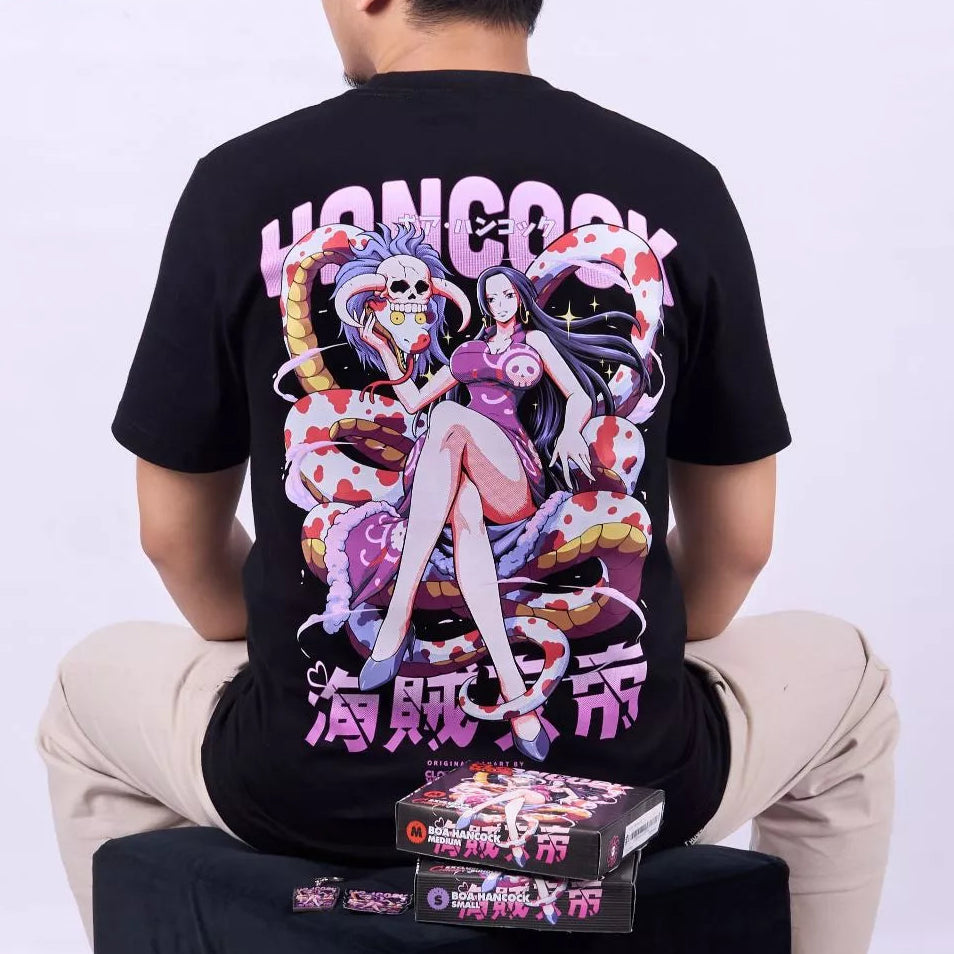 A man wearing a black graphic T-shirt from the back, seated against a light gray studio background. The shirt features a large anime-style print with pink, purple, white, and red tones, plus large pink text.